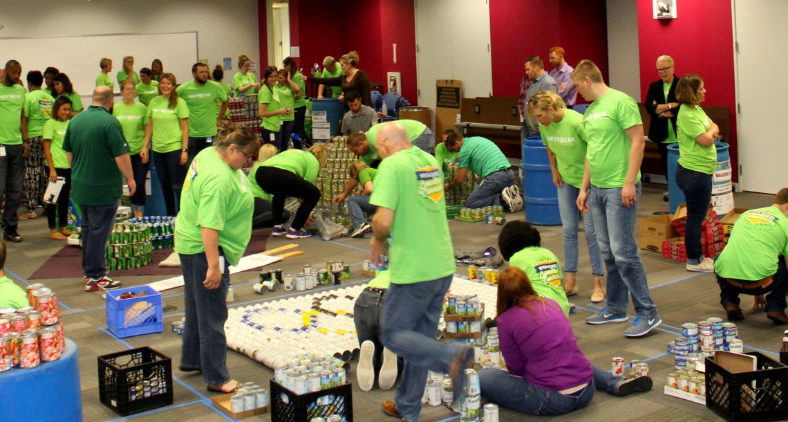 CANStruction volunteers in green matching shirts work together to build structures out of canned goods